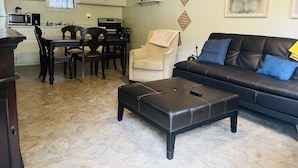 Una sala de estar con sofá, sillón y reposapiés. Hay una mesa de comedor con sillas y una zona de cocina con armarios y electrodomésticos.