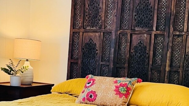 Una cama con colcha amarilla y almohadas florales, cabecera de madera con detalles tallados y una mesita de noche con una lámpara y una planta.