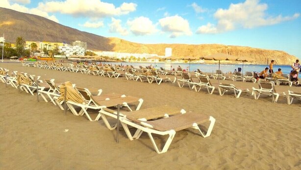 A beach with rows of sun loungers and umbrellas, a clear sky, and a mountain in the background.