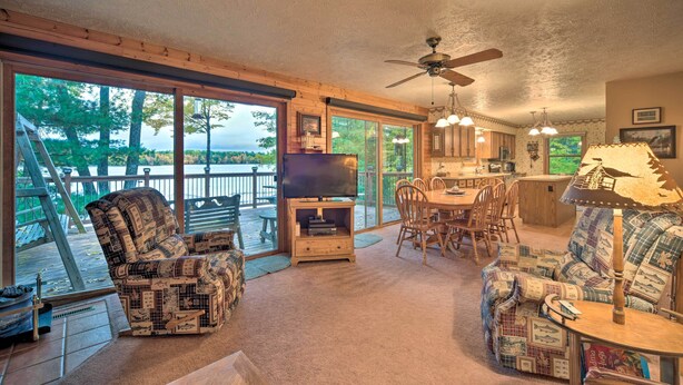 A cozy living room with a television, wooden furniture, and large windows overlooking a deck. - Cozy Hayward Cottage w/ Dock & Lakefront View (Hayward)