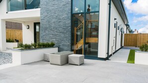 A modern outdoor patio with a stone floor, two grey wicker sofas, and a wooden table. - Gwel y Wyddfa - sleeps 6 guests in 3 bedrooms (Rhosneigr)