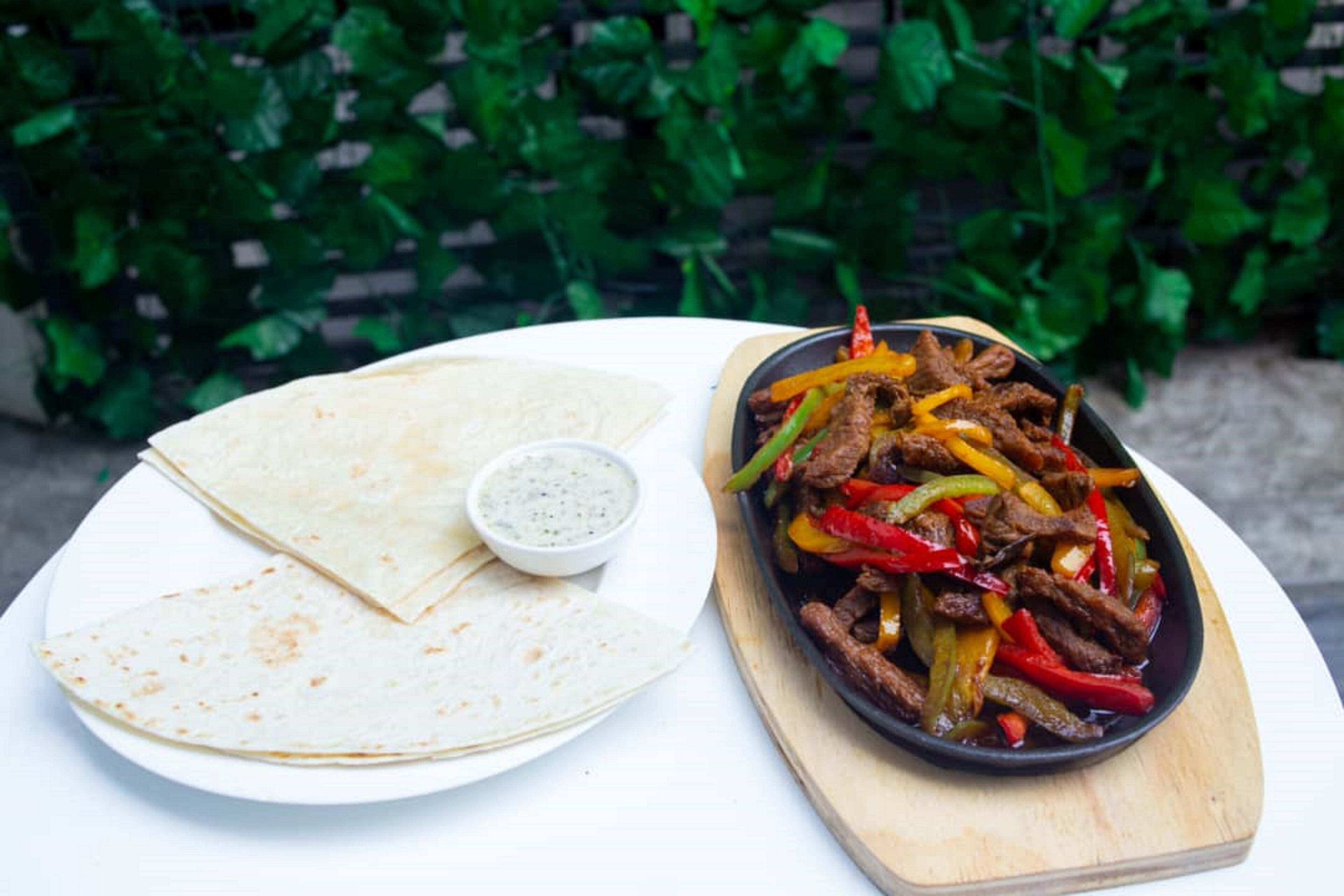 A plate of flatbread, a bowl of sauce, and a skillet with beef and bell peppers.