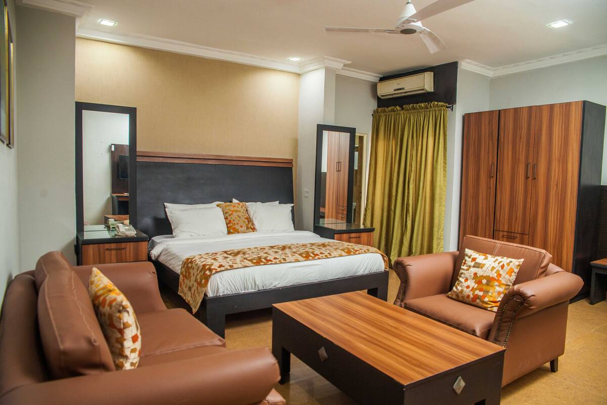 Una habitación de hotel con cama, dos sofás, una mesa de centro de madera, armario y ventilador de techo.