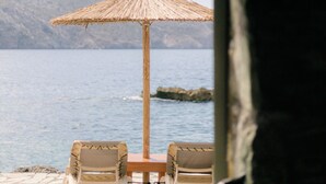 Blick auf einen Tisch und Stühle am Strand, mit einem Strohdachschirm, mit Blick auf ein ruhiges Meer und ferne Berge.