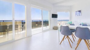 Un'ampia stanza luminosa con grandi finestre, un'area pranzo con sedie e un televisore a schermo piatto appeso al muro.