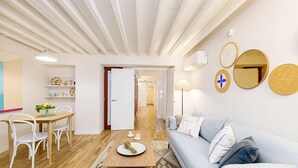 Una sala de estar moderna con suelo de madera, un sofá azul claro, sillas blancas, una mesa de comedor de madera y una zona de cocina al fondo.