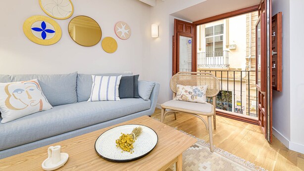 Una sala de estar acogedora con un sofá, una mesa de comedor con un plato de comida y una silla. Hay platos decorativos en la pared y un balcón con barandilla.