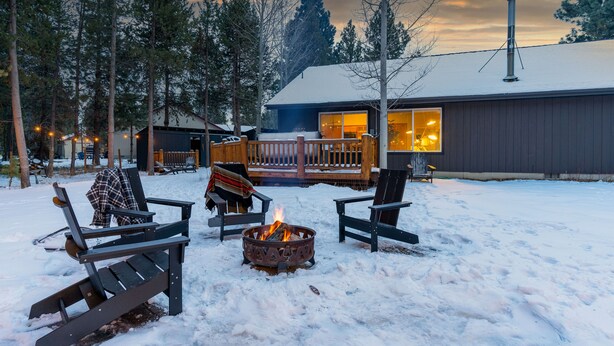 A snow-covered deck with Adirondack chairs and a fire pit, surrounded by a wooden deck and a building with large windows. - Bend Modern Cabin w/ Private Hot Tub . Fenced Yard, Game Room, Pet-Friendly (Bend)