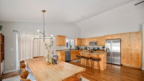 A spacious kitchen with wooden cabinets, a large island with bar stools, a stainless steel refrigerator, and a dining area with a wooden table and chairs. - Bend Modern Cabin w/ Private Hot Tub . Fenced Yard, Game Room, Pet-Friendly (Bend)