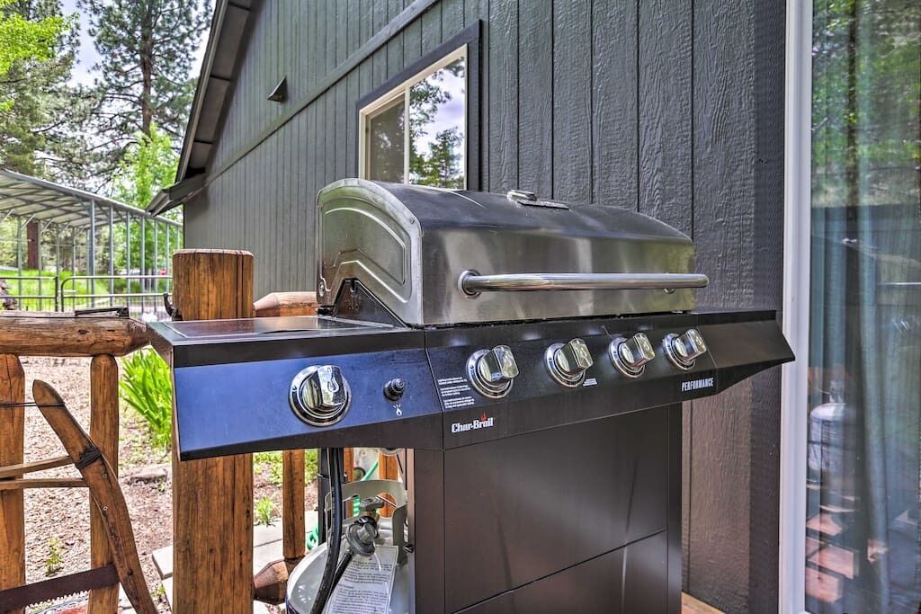 A stainless steel Char-Broil grill with four burners and a chimney, positioned outdoors next to a wooden deck and a house with a window.
