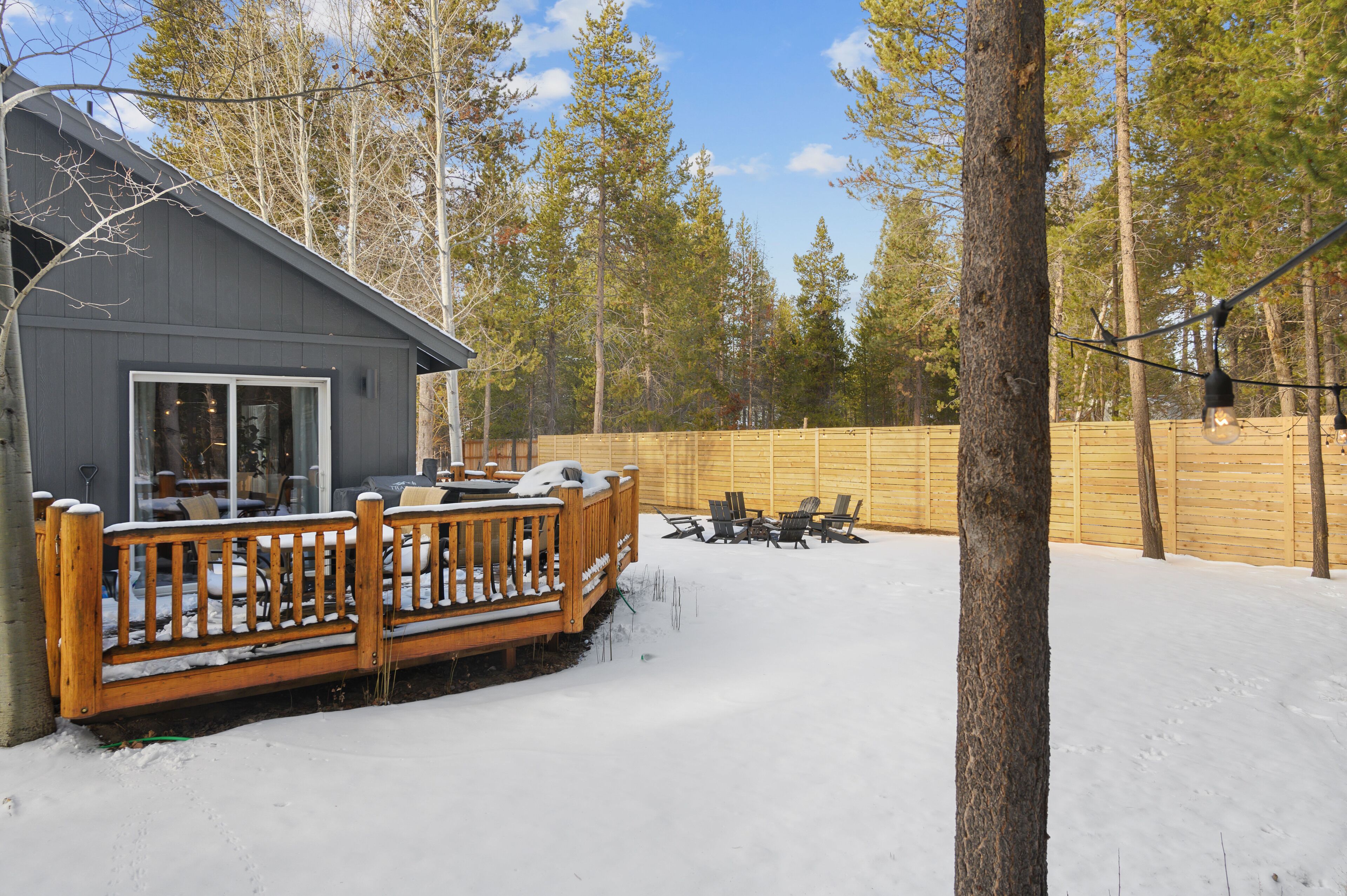 A wooden deck with chairs and a table, a building with a large window, and a wooden fence in a snowy outdoor setting.