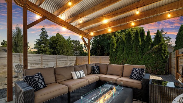 Un patio exterior con una pérgola de madera, un pozo de fuego con tapa de vidrio y un sofá seccional marrón con cojines estampados.