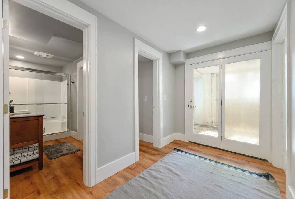 A spacious hallway with a wooden floor, a grey rug, and a white door leading to a bathroom with a glass shower.