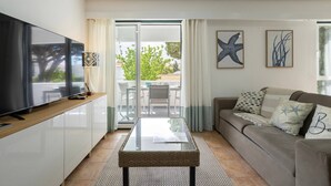 A modern living room with a flat-screen TV, a glass-top coffee table, a sofa with patterned cushions, and a balcony with a view. - Amazing Apt. in the Heart of Vilm by Centralgarve (Quarteira)