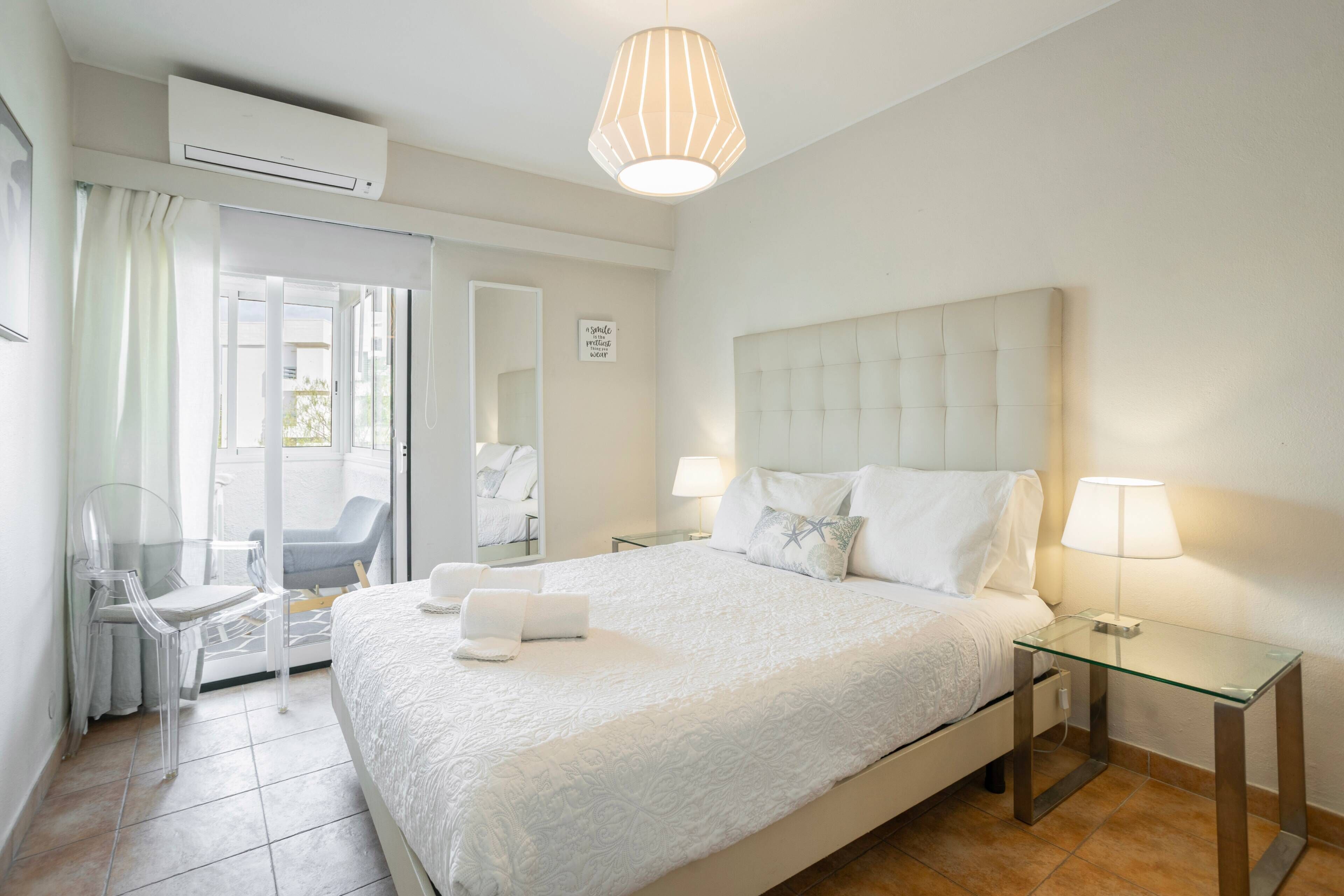 A hotel room with a bed, two bedside tables, a glass table, a chair, a mirror, and an air conditioning unit.