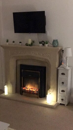 Ein Kamin mit darüber montiertem Fernseher, umgeben von Dekoration und einer kleinen Kommode mit einer Lampe.
