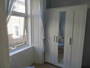 Ein Zimmer mit Fenster, weißem Kleiderschrank und einem Bett mit gestreifter Bettdecke.