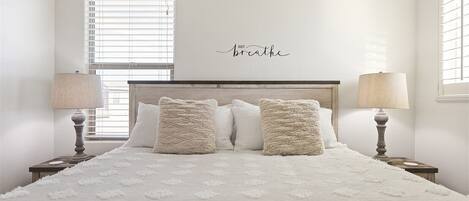 A bedroom with a large bed, two bedside lamps, and a wall-mounted sign that says "Just breathe".