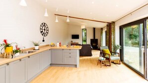 A modern kitchen with a central island, grey cabinets, and a wooden countertop. There is a dining area with a sofa and a chair, and a large window providing a view of the outdoors.