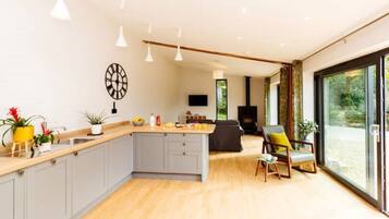 Une cuisine moderne dotée d’un îlot central, de meubles gris et d’un plan de travail en bois. On y trouve un coin repas avec un canapé et un fauteuil, ainsi qu’une grande fenêtre offrant une vue sur l’extérieur.