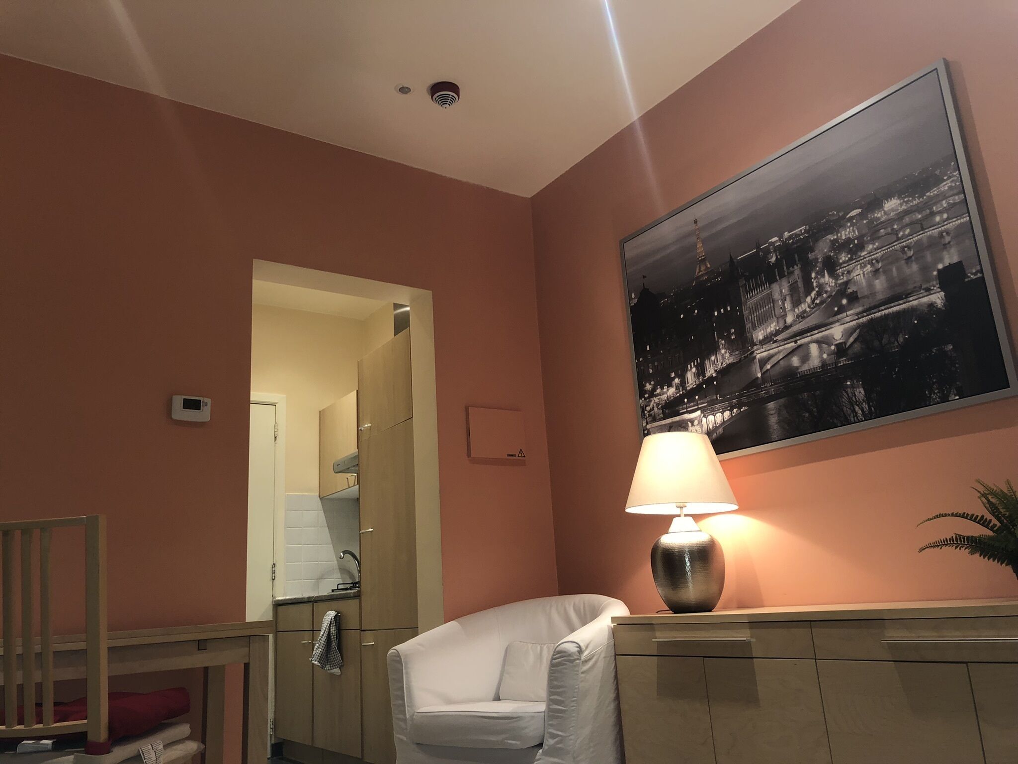 A hotel room with a white chair, a night lamp, a framed cityscape picture, and a wooden cabinet.