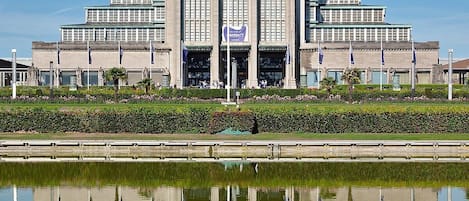 Um edifício grande e simétrico com uma entrada central, ladeado por estátuas e bandeiras, refletido em águas calmas.