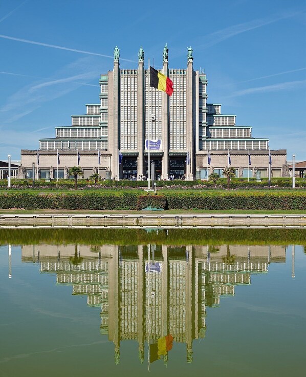 A large, symmetrical building with a central entrance, flanked by statues and flags, reflecting in a calm body of water.
