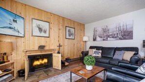 Una sala de estar acogedora con chimenea, un sofá de cuero negro, una mesa de centro y un televisor montado.