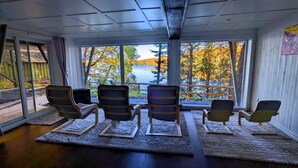 Ein Zimmer mit fünf Stühlen vor einem großen Fenster mit Blick auf einen See und Herbstbäume.