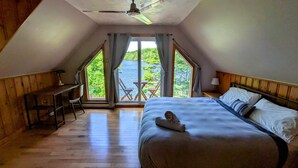 Ein Schlafzimmer mit einem großen Bett, einem Schreibtisch und einem Stuhl. Ein Fenster mit Vorhängen bietet Blick auf viel Grün.