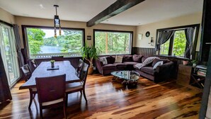 Ein geräumiges Wohnzimmer mit Holzboden, einem Esstisch mit Stühlen, einer Couch und einem Couchtisch. Große Fenster bieten Blick auf Grünflächen und ein Gewässer.