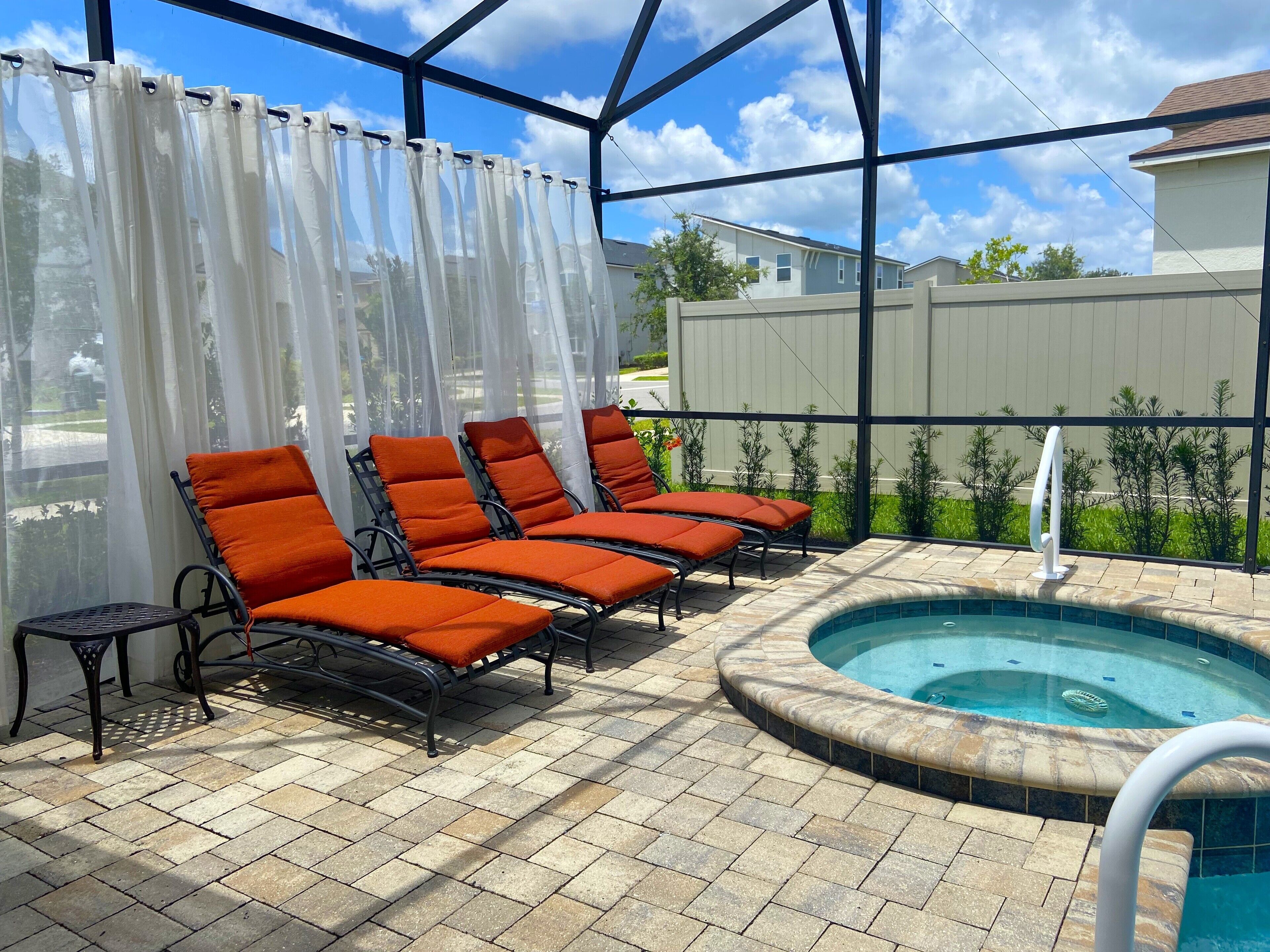 A covered outdoor area with a hot tub, lounge chairs, and a view of a residential neighborhood.