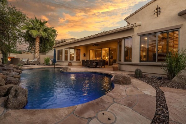 A pool with a stone border, a patio with outdoor furniture, and a building with large windows and a covered patio area. - Gilbert 4BR Villa|Heated Pool+Games|Sleeps 12 (Gilbert)