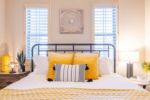 Une chambre à coucher avec un lit, deux fenêtres, une table de chevet, une lampe et un élément décoratif mural.