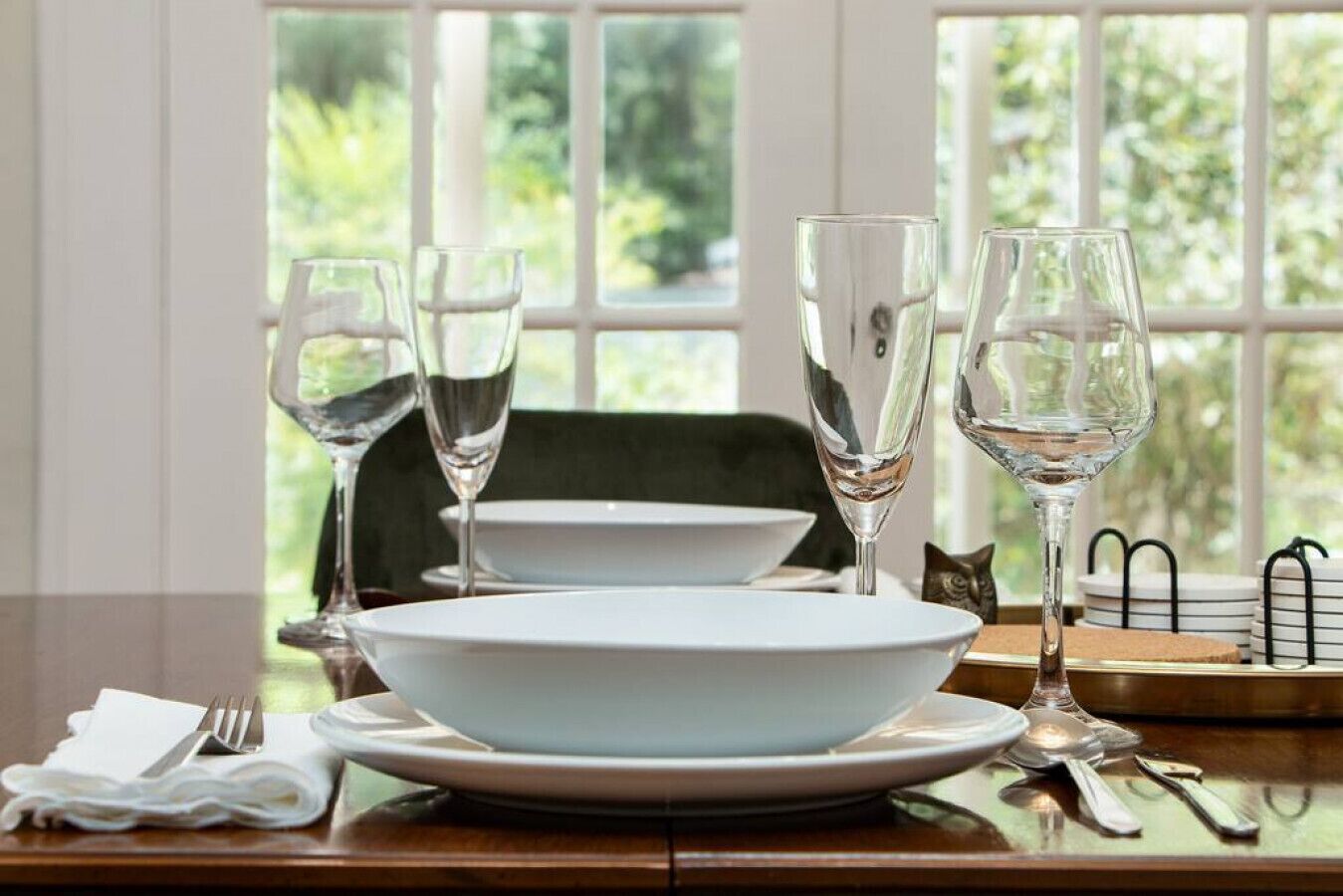 Une table à manger dressée avec de l’argenterie blanche, des verres transparents et des couverts, offrant une vue sur la verdure par la fenêtre.