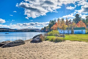A lakeside scene with a sandy beach, a wooden bench, and a small cabin with a green roof. - Fire Pit & Beach: Lake Pleasant Home w/ Views (Speculator)