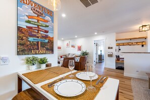 A dining area with a large poster of Utah landmarks, a modern kitchen, and a living room with a brown leather sofa. - 2 Bedroom Near Zion•Bryce•Wave: Hot Tub, Dining/Patio & Games - Red Rock Views! (Kanab)