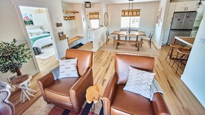 Un vaste salon agrémenté d’une grande fenêtre, un coin repas avec une table et des chaises en bois, et une cuisine équipée d’appareils électroménagers modernes.