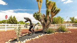 Un panneau de signalisation indiquant un arrêt obligatoire, un cactus et une sculpture métallique représentant un dinosaure, le tout situé dans un espace aménagé avec une clôture blanche et sous un ciel dégagé.