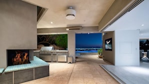 A modern outdoor kitchen with a stone countertop, a built-in grill, and a seating area with a view of the city at night. - Camelback Vista - 4BR Luxury Estate w/ Infinity Pool & Camelback Views (Phoenix)