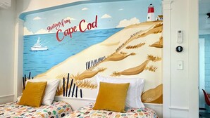 A bedroom with a mural of Cape Cod, a bed with patterned bedding, and a nightstand with a lamp.