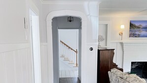 A hallway with a white door leading to a staircase, a wooden desk, a patterned sofa, and a fireplace with a painting above it.