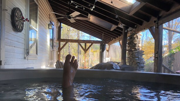Un jacuzzi con una mano en el agua, vigas de madera, un ventilador de techo y un dardo en la pared.