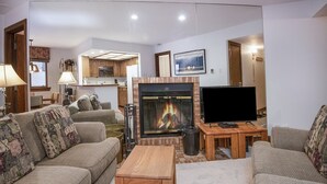 A living room with a fireplace, sofas, a television, and a kitchen in the background. - Village Inn Plaza 103 (Vail)