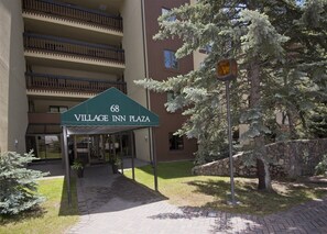 The entrance to Village Inn Plaza, featuring a green awning with the address and name, a stone bridge, and a tree in the foreground. - Village Inn Plaza 103 (Vail)