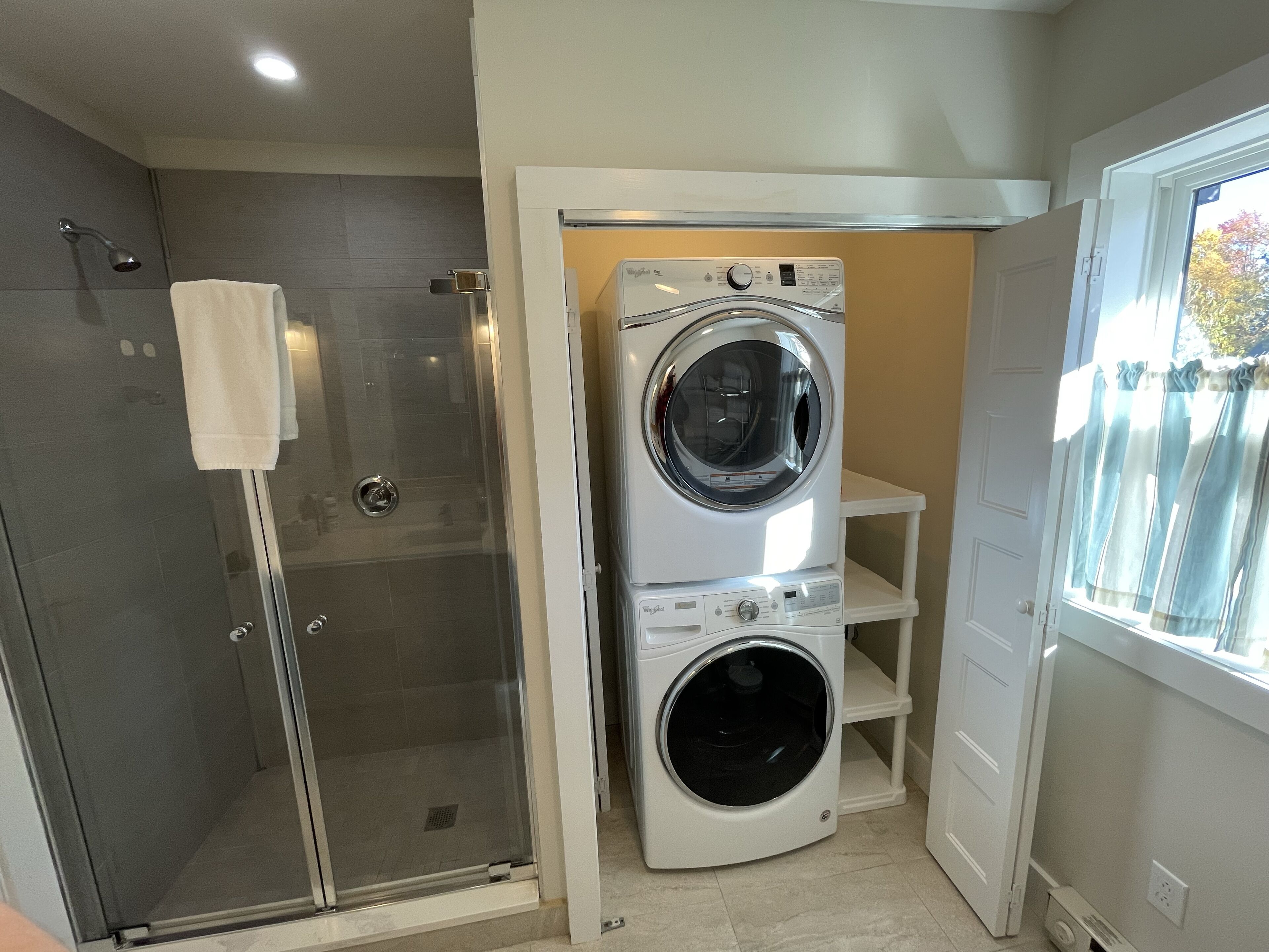 A compact laundry room with a front-loading washer and dryer, a glass shower enclosure, and a small window with curtains.
