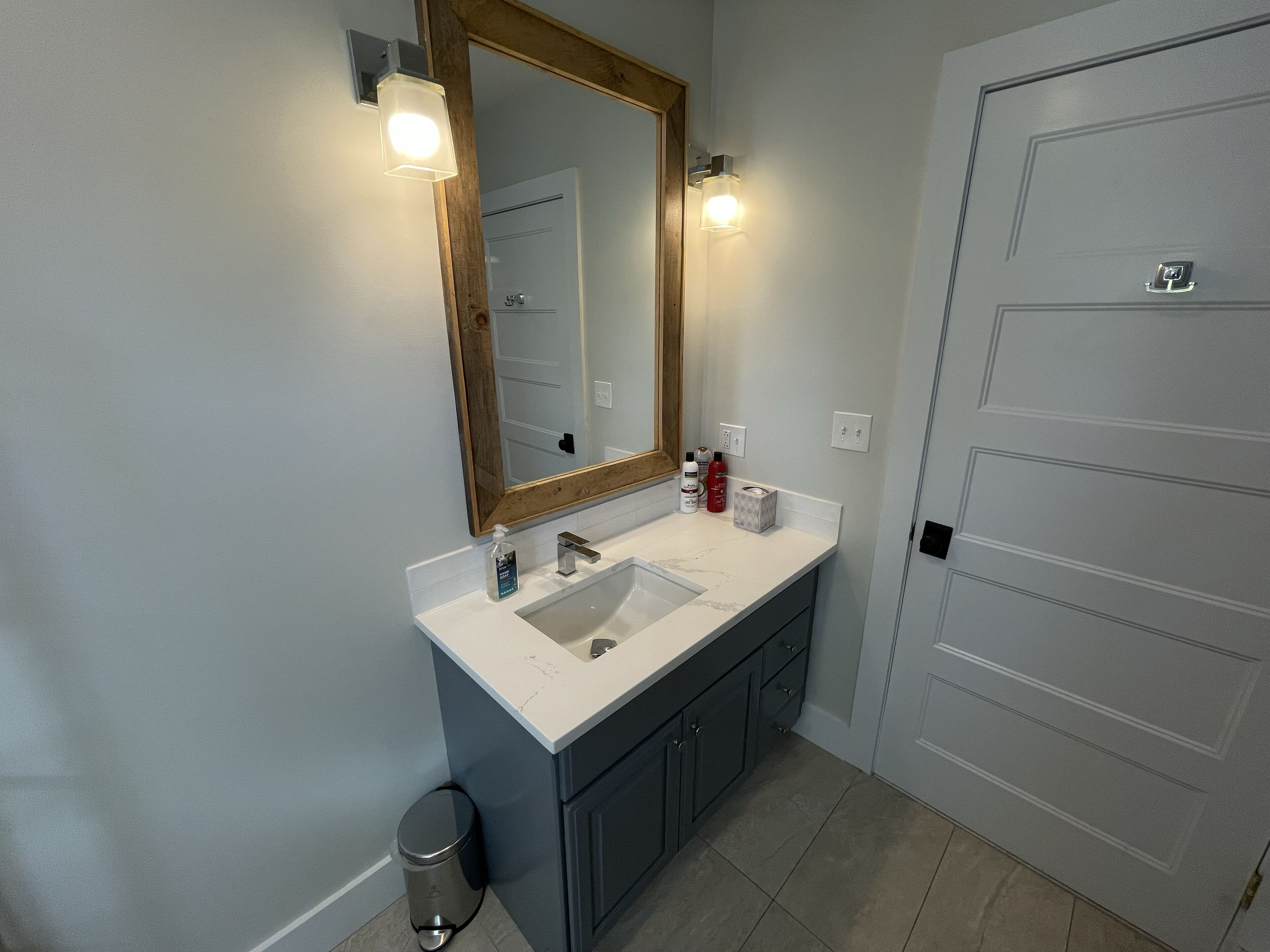 Un baño con lavamanos blanco, muebles oscuros, un espejo con iluminación y una puerta con picaporte.