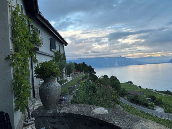 Una balaustra in pietra con una pianta in vaso, un edificio ricoperto di edera e vista su un lago con montagne sullo sfondo.