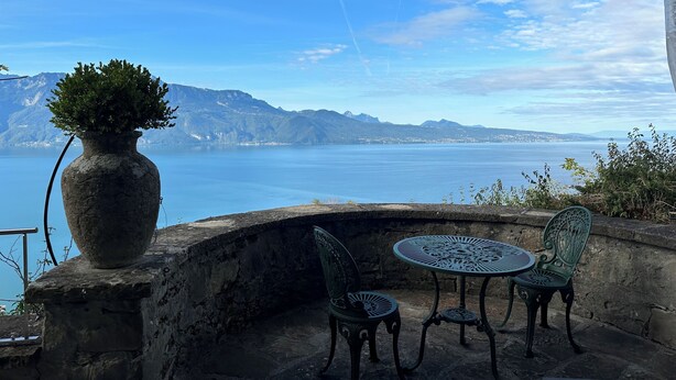 Un espacio al aire libre con una mesa y dos sillas, un jarrón grande con una planta, y vistas a un lago con montañas al fondo.