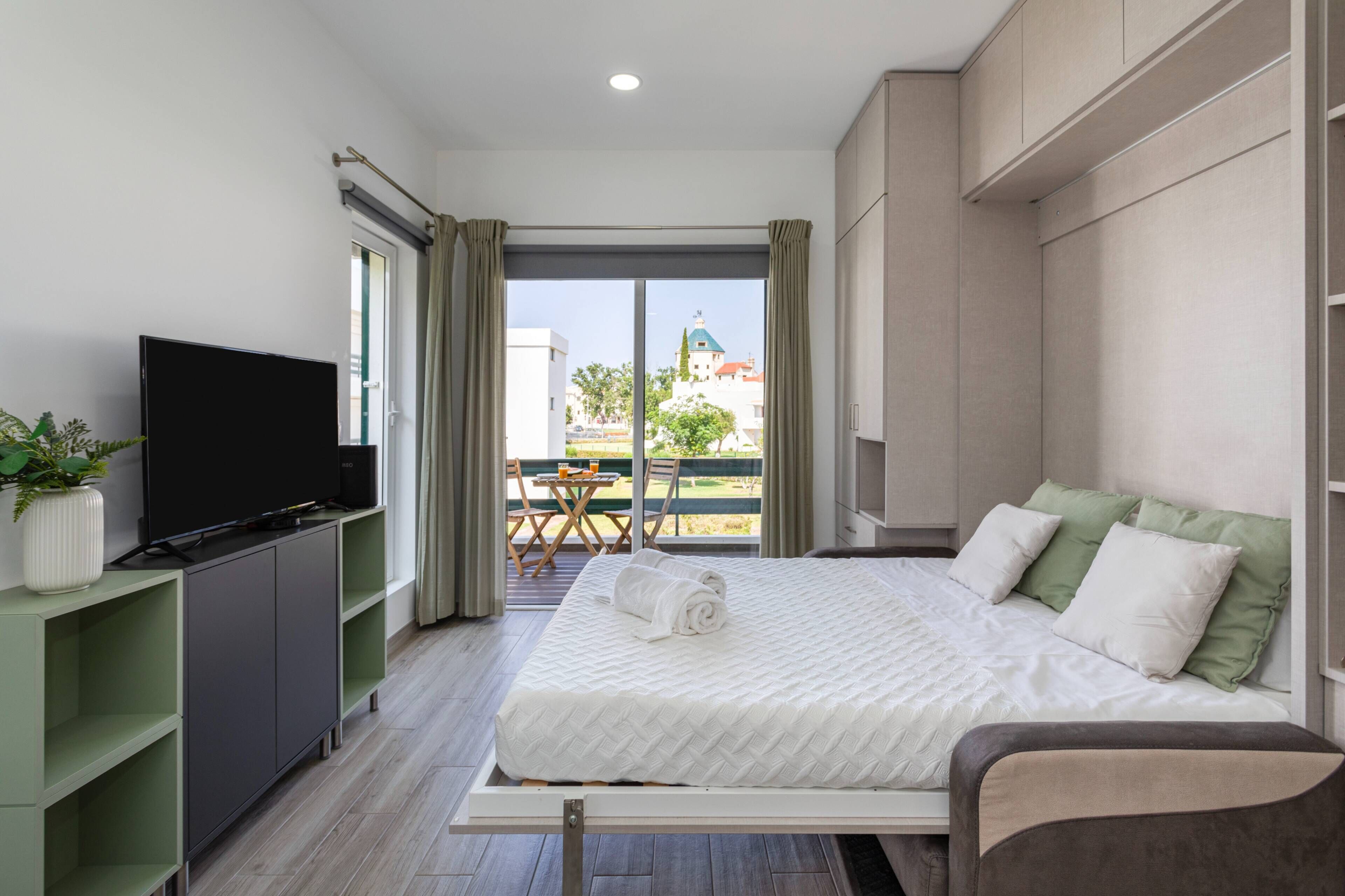 Een moderne hotelkamer met een groot bed, een flatscreen-tv, een groen dressoir en een balkon met een tafel en stoelen.